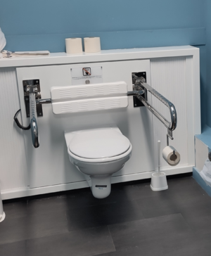 An einer hellblauen Wand ist ein weißer, rechteckiger Kasten montiert, der bis zum Boden reicht. Am dessen oberer Innenkante befindet sich mittig ein weiterer schmalerer weißer Kasten, an dem Stützklappgriffe (mit Klorollenhalter rechts bei Draufsicht) und die bewegliche Rückenlehne geschraubt sind.
